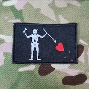 May include: Black fabric patch with a white skeleton holding a spear and a red heart with three red dots below it.