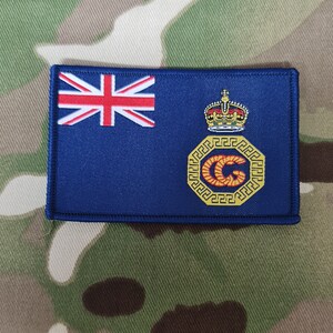 Puede incluir: Un parche bordado azul con una corona dorada y un círculo dorado con una "G" roja en su interior. El parche también presenta una pequeña bandera del Reino Unido en la esquina superior izquierda.