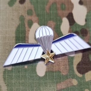 Op de afbeelding: Een witte en blauwe emaille pin met een parachute en vleugels. De parachute is zilver en de vleugels hebben een blauwe streep. Een gouden ster bevindt zich in het midden van de vleugels.