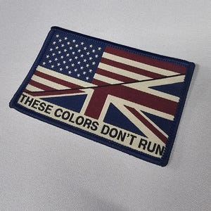 Op de afbeelding: Rechthoekige patch met een combinatie van de Amerikaanse en Britse vlaggen. De bovenste helft toont de Amerikaanse vlag, de onderste helft de Union Jack. De tekst "THESE COLORS DON'T RUN" is in het wit gedrukt.