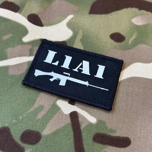 Può includere: Toppa rettangolare nera con testo bianco "LIAI" e grafica di un fucile bianco. La toppa è posta su uno sfondo con motivo mimetico.