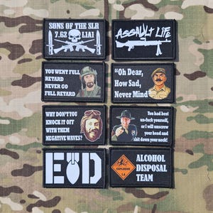 Puede incluir: Una colección de ocho parches rectangulares negros con varios textos blancos y diseños gráficos, incluyendo ilustraciones militares, citas de películas y eslóganes humorísticos, dispuestos en dos columnas sobre un fondo de camuflaje.