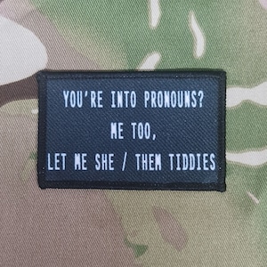 Può includere: Toppa nera con testo bianco che dice "You're into pronouns? Me too, let me she/them tiddies."