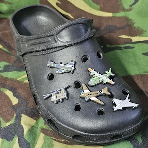 Peut inclure: Sabot noir orné de breloques en forme d'avions. Les breloques représentent un hélicoptère, un avion de chasse et plusieurs avions à hélices de différentes couleurs. La chaussure repose sur un fond de camouflage.