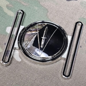 May include: A shiny silver belt buckle with a circular center featuring a knife design. The buckle has two parallel bars on either side. The background is a camouflage pattern.