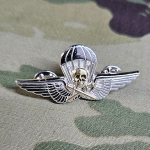 May include: Silver and gold skull and crossbones with a parachute above the skull. The wings are silver and have a black outline. The wings are spread out and the parachute is above the skull. The pin is on a camouflage background.