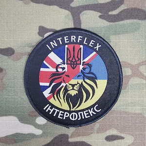 Può includere: Una toppa rotonda nera con la scritta "INTERFLEX" in bianco. La toppa presenta un disegno di leone con le bandiere del Regno Unito e dell'Ucraina. Lo sfondo è mimetico.