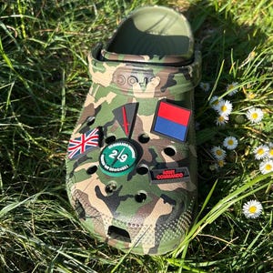 Puede incluir: Zuecos con estampado de camuflaje con varios abalorios para zapatos de temática militar. Los abalorios incluyen una bandera roja, blanca y azul, una bandera negra y roja, un círculo verde con el texto "29 Commando Brigade" y el texto "United We Conquer", y un abalorio negro con el texto "Army Commando" en rojo.