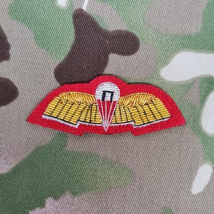 May include: A red and gold embroidered paratrooper badge with a white parachute and a black 'H' inside. The badge is on a camouflage fabric background.