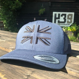 Puede incluir: Una gorra de béisbol azul y negra con un diseño de Union Jack bordado en negro. La gorra tiene una parte trasera de malla y un cierre a presión.