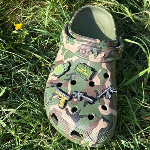 May include: Clogs with a camouflage pattern featuring various military-themed charms, including a helmet, a gun, and a sign that reads "Front Toward Enemy."