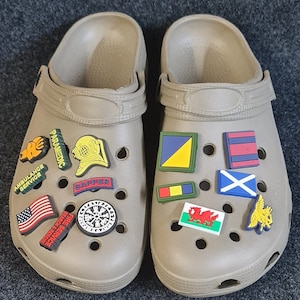 May include: A pair of tan clogs with various colorful charms attached. The charms include a US flag, a fire service emblem, a compass, a paramedic symbol, a diver's helmet, a "sapper" patch, a Welsh dragon, a Scottish flag, a nautical flag, and a golden eagle.