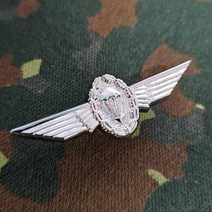 May include: A silver-colored military paratrooper badge with a parachute design in the center, surrounded by a wreath. The badge has two wings extending outward. The background is a camouflage pattern.