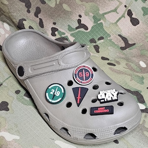 Könnte beinhalten: Grauer Clog-Schuh mit mehreren dekorativen Ansteckern. Die Anstecker zeigen Text und Grafiken wie "COMMANDO SQUADRON ROYAL ENGINEERS", "2/9", ein rotes Schwert und "SOUNDS GAY I'M IN".
