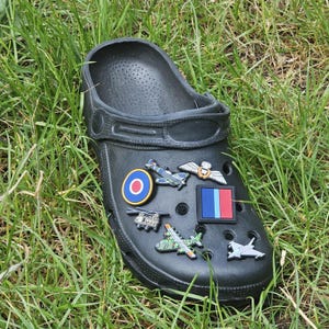 May include: Black clog shoe decorated with various aviation-themed charms. The charms include miniature airplane, helicopter, and emblem designs in shades of blue, green, white, and gray. The shoe is set against a backdrop of green grass.