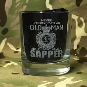 May include: A clear glass tumbler with a black interior. The tumbler features the text "NEVER UNDERESTIMATE AN OLD MAN WHO IS ALSO A SAPPER" in silver lettering, along with a crest. The glass is on a camouflage background.