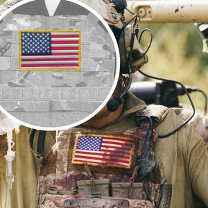 Può includere: Un soldato indossa un giubbotto mimetico con due toppe della bandiera americana. Il giubbotto ha più tasche e cinghie. Il soldato indossa un auricolare e ha un microfono attaccato al petto.
