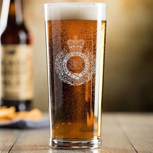 May include: A pint glass filled with beer, featuring a Royal Engineers emblem. The emblem includes a crown, the initials "ER", and the motto "Honi Soit Qui Mal Y Pense". The glass is on a wooden surface, with a blurred bottle in the background.