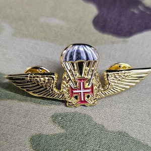 May include: A gold-colored pin with a red and white cross, a parachute, and wings. The pin is a symbol of the Portuguese Air Force.