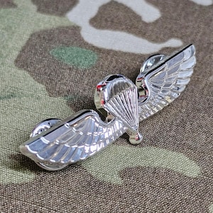 May include: Silver-toned military paratrooper wings with a parachute design. The wings are on a camouflage fabric background.