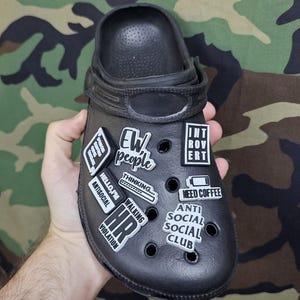 May include: Black Crocs shoe with various white and black charms attached. Charms feature phrases such as "EW people", "Introvert", "Need Coffee", and "Anti Social Social Club". The shoe is held in a hand.