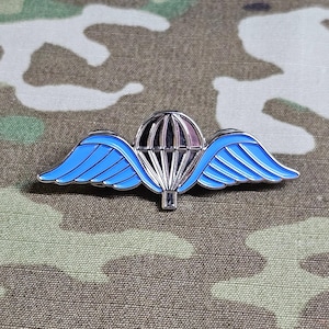 May include: A silver-colored pin featuring a parachute design with light blue wings. The parachute is detailed with black and silver lines, and the wings have a blue enamel finish. The pin is set against a camouflage background.