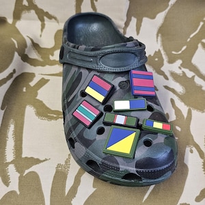 May include: A green camouflage clog shoe decorated with a variety of rectangular charms. The charms display striped and geometric designs in red, blue, yellow, and green. The shoe is placed on a camouflage patterned surface.