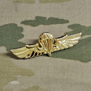 Pode incluir: Um distintivo de paraquedista militar dourado com asas e um paraquedas. O distintivo está sobre um fundo de tecido de camuflagem.