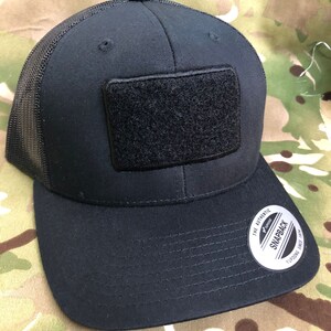 Può includere: Cappellino da camionista nero in rete con una toppa in velcro nera sul davanti. Il cappello ha una visiera nera e una chiusura a scatto nera. Il cappello è etichettato "The Authentic Yupoong Snapback Since 1974".