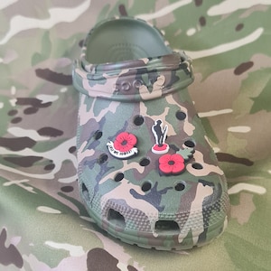 Może przedstawiać: Klapki w kamuflażowym wzorze z trzema czerwonymi charmsami w kształcie maków, jeden z nich z napisem "Lest We Forget".