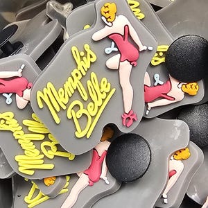 May include: A collection of grey shoe charms with a pin-up girl design and the text "Memphis Belle" in yellow. The pin-up girl wears a pink dress and heels. Black round charms are also visible.