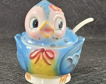 May include: A blue and yellow ceramic bird sugar bowl with a red bow and a white spoon. The bird has a pink face with large eyes and a yellow beak.