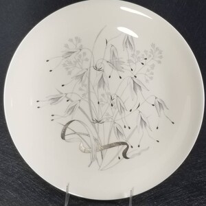 May include: A white ceramic dinner plate with a silver floral design. The design features a bouquet of stylized flowers and leaves.