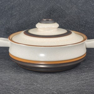 May include: A white ceramic casserole dish with a brown and black striped lid. The dish has two handles and a brown and orange rim.