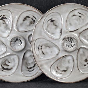 May include: Two ceramic oyster plates. Each plate is round, with six oyster-shaped indentations and a central well. The plates are a mottled gray with dark brown edges and accents. The plates are designed for serving oysters.
