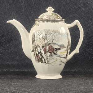 May include: A white ceramic teapot with a winter scene design. The teapot has a brown handle and a lid with a floral design. The scene depicts a red barn in a snowy landscape with trees and a fence.