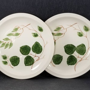 May include: Two white ceramic dinner plates with a green leaf design. The leaves are painted in a light green and dark green color.