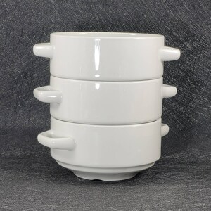 May include: Three white ceramic bowls with handles stacked on top of each other. The bowls are round and have a smooth, glossy finish.