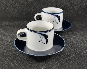 May include: Two white ceramic coffee cups with blue and black floral designs on a dark blue saucer.