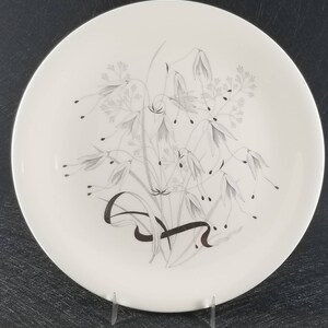 May include: A white ceramic dinner plate with a silver and gray floral design. The design features a cluster of stylized flowers and leaves.