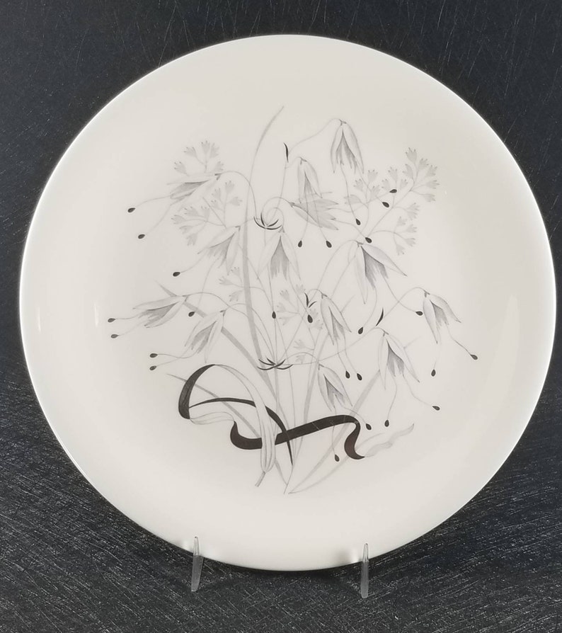 May include: A white ceramic dinner plate with a silver and gray floral design. The design features a cluster of stylized flowers and leaves.