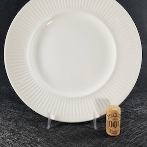 May include: A white ceramic dinner plate featuring a smooth centre and a wide rim decorated with vertical ribbed detailing. The plate is displayed upright on a clear stand next to a small cylindrical cork stopper with the text 100 printed on it.