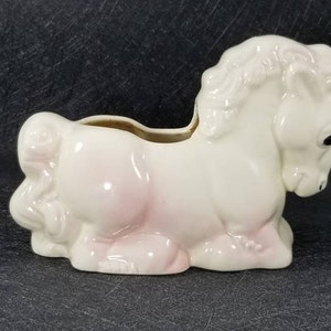 May include: White ceramic planter shaped like a lying down horse with pink accents. The planter has a small opening on the back for planting.