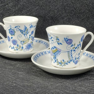 May include: Two white teacups with blue and green floral patterns and matching saucers. The cups have a stylized bird and a stylized woman design.