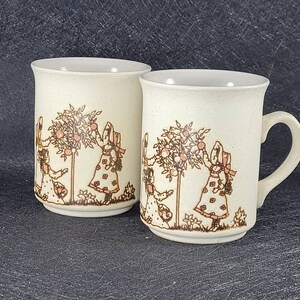 May include: Two ceramic mugs with a cream-coloured speckled finish. Each mug features a brown illustration of children picking fruit from a tree. The mugs have a white interior and a curved handle. Suitable for hot or cold drinks.
