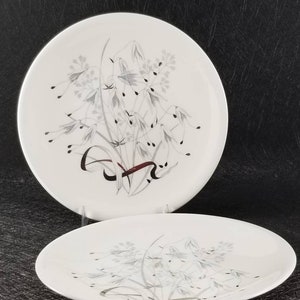 May include: Two white ceramic dinner plates with a silver and gray floral design. The design features a cluster of stylized flowers and leaves with a red ribbon.