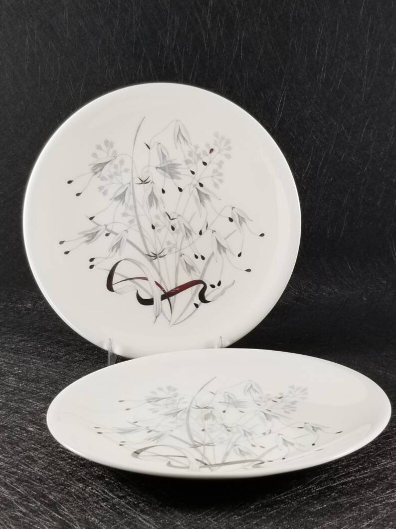 May include: Two white ceramic dinner plates with a silver and gray floral design. The design features a cluster of stylized flowers and leaves with a red ribbon.