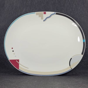 May include: A white oval serving platter with a geometric design in shades of blue, red, and beige. The design features a wavy line, a rectangle, and a circle.