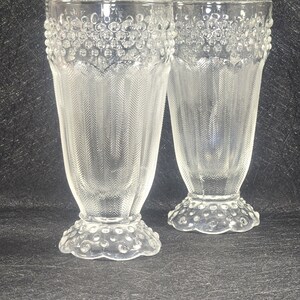 May include: Two clear, vintage-style drinking glasses. The glasses have a flared shape with a textured, bubble-like design at the rim and base. The body of the glasses features a vertical, herringbone pattern. The glasses are likely made of glass.