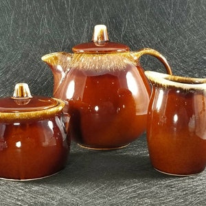 May include: A brown glazed ceramic teapot, creamer and sugar bowl set. The teapot has a lid with a knob on top. The creamer and sugar bowl have lids.